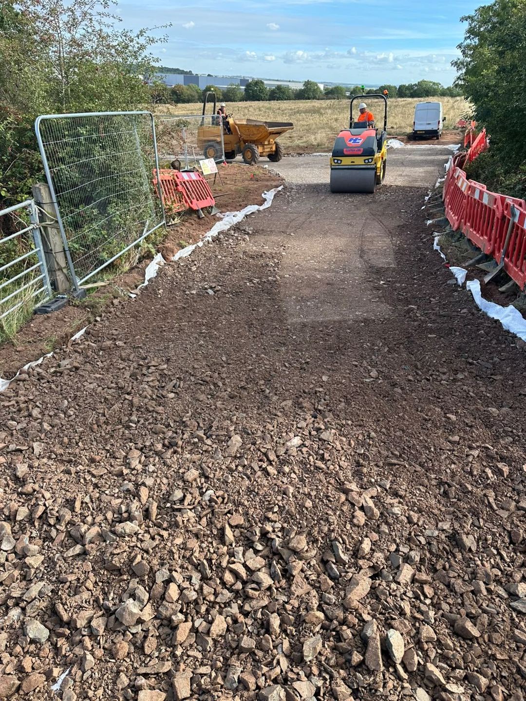 A roller compacting crushed stone on a fenced access track, with a dumper working further along.