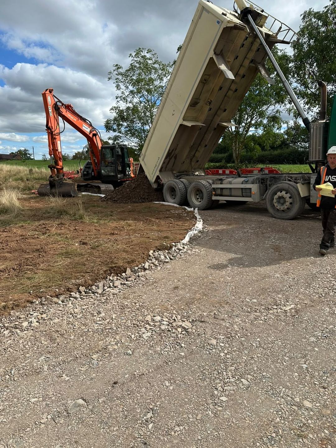A tipper lorry discharging crushed stone onto a new access track as an excavator works behind.