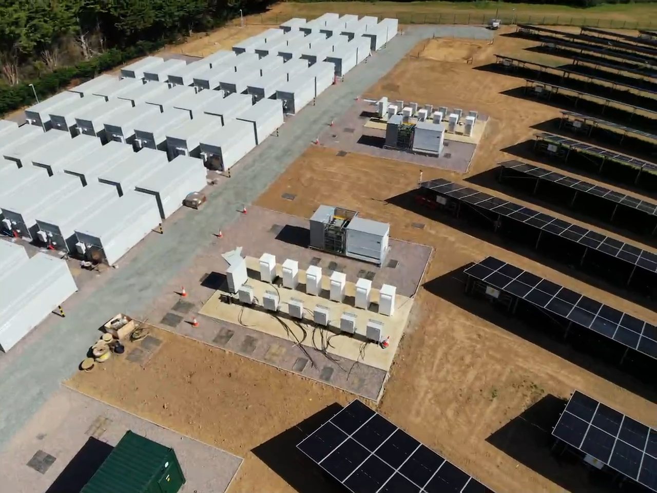 Aerial view of battery storage containers, inverters and transformers on their bases beside solar panel rows.