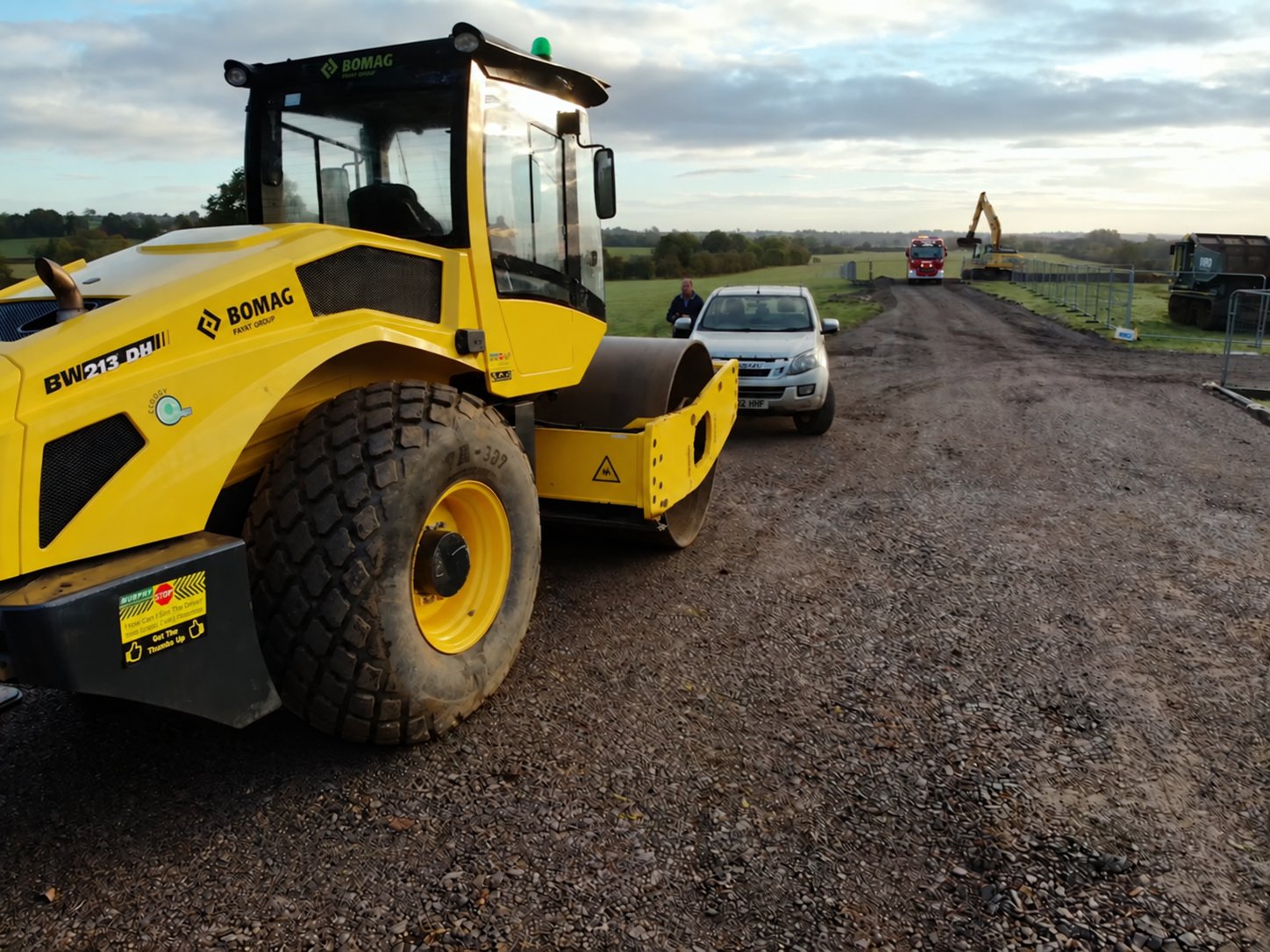 A ride-on roller compacting the stone surface of a newly built haul road, with an excavator working in the distance.