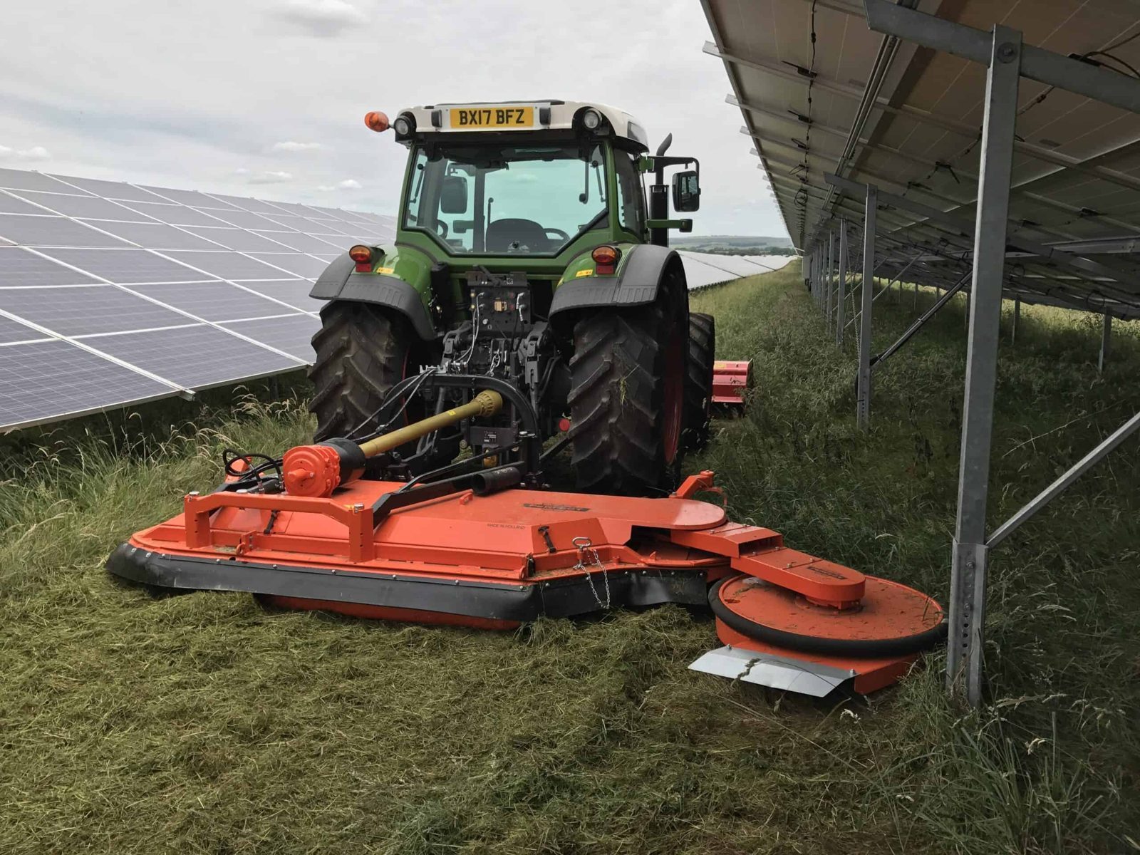 A tractor with a mounted mower cutting vegetation between solar panel rows during grounds maintenance at a solar farm.