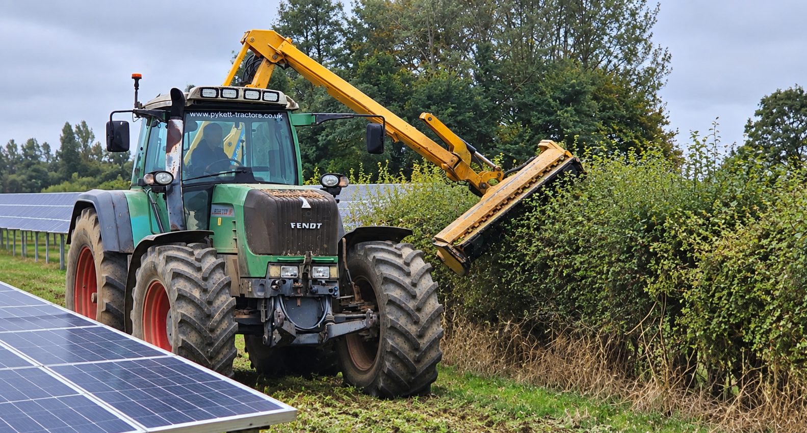 A tractor with a reach-arm flail trimming a boundary hedge beside the panel rows at a solar farm.
